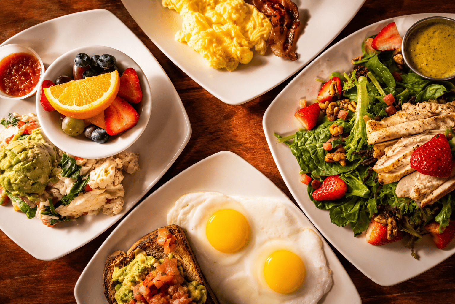 A protein-forward spread — egg white scramble with avocado, fresh fruit, eggs with avocado toast, and a grilled chicken strawberry salad